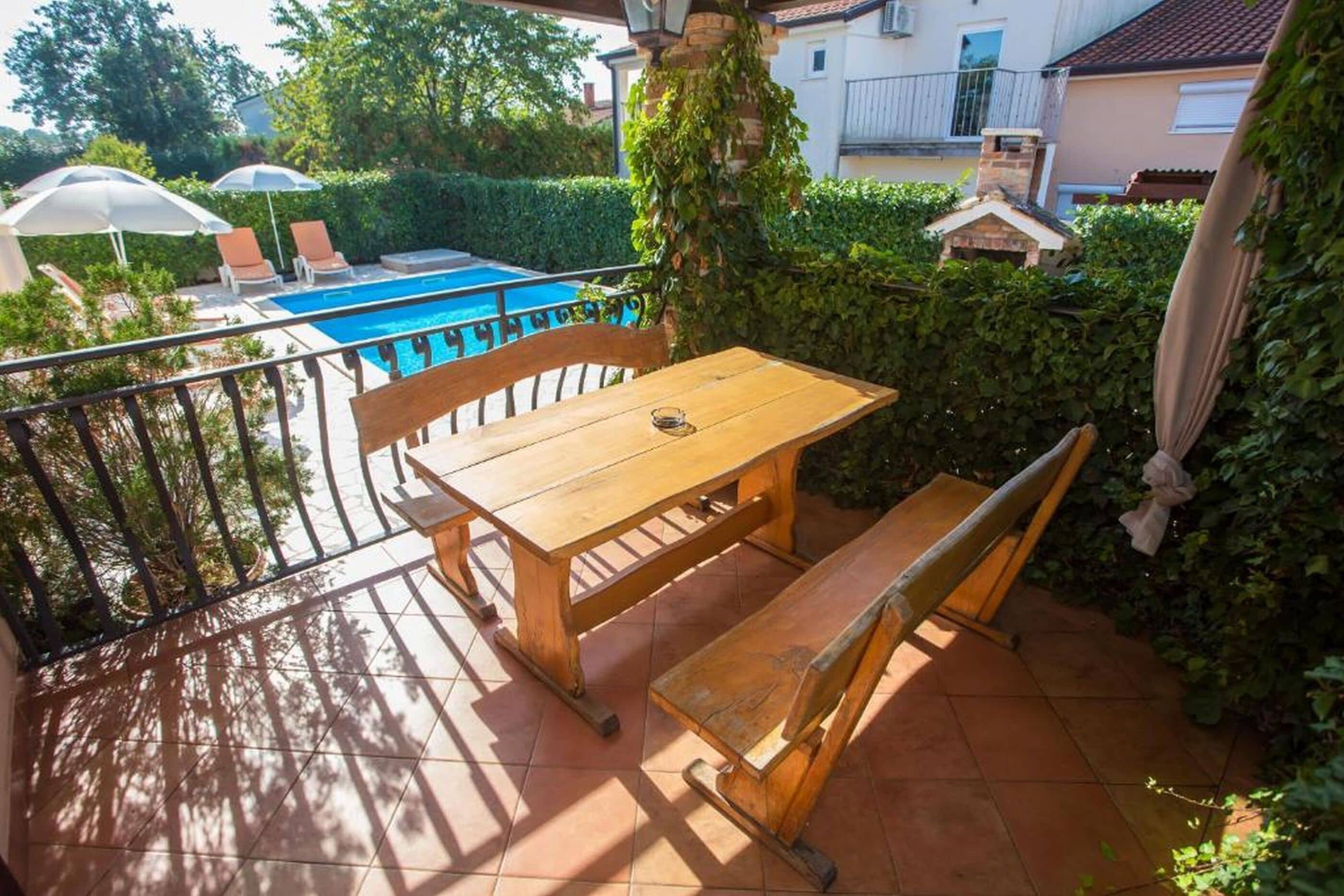 A wooden table and bench on a balcony with a pool in the background.