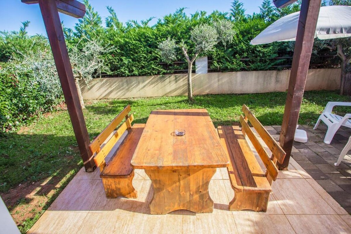 Ein hölzernes Picknicktisch mit Bänken, umgeben von einem Garten mit Hecke und Baum.