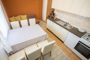 A compact kitchenette with a stove, sink, and a small countertop. There is a bed with a patterned bedspread and two chairs. - Apartments Eny (Porec)