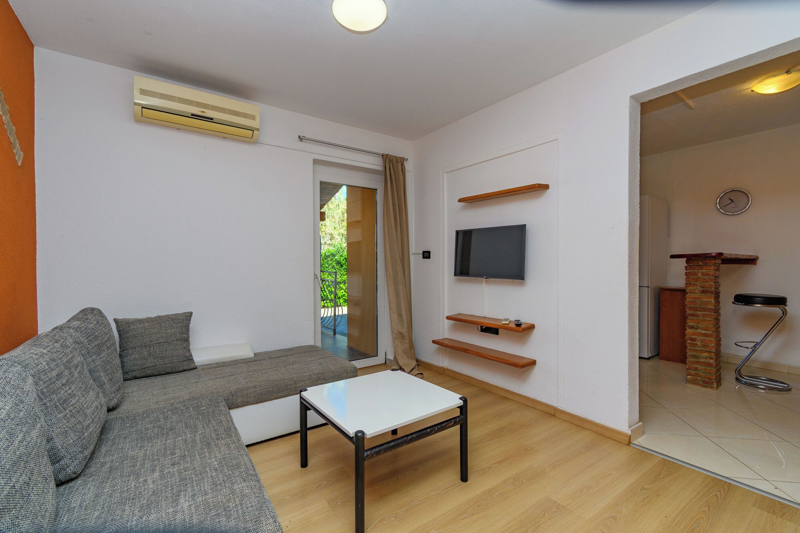 Una sala de estar con un sofá gris, una mesa de centro blanca, un televisor de pantalla plana montado en la pared y un puerta corredera de vidrio que da a un espacio exterior.