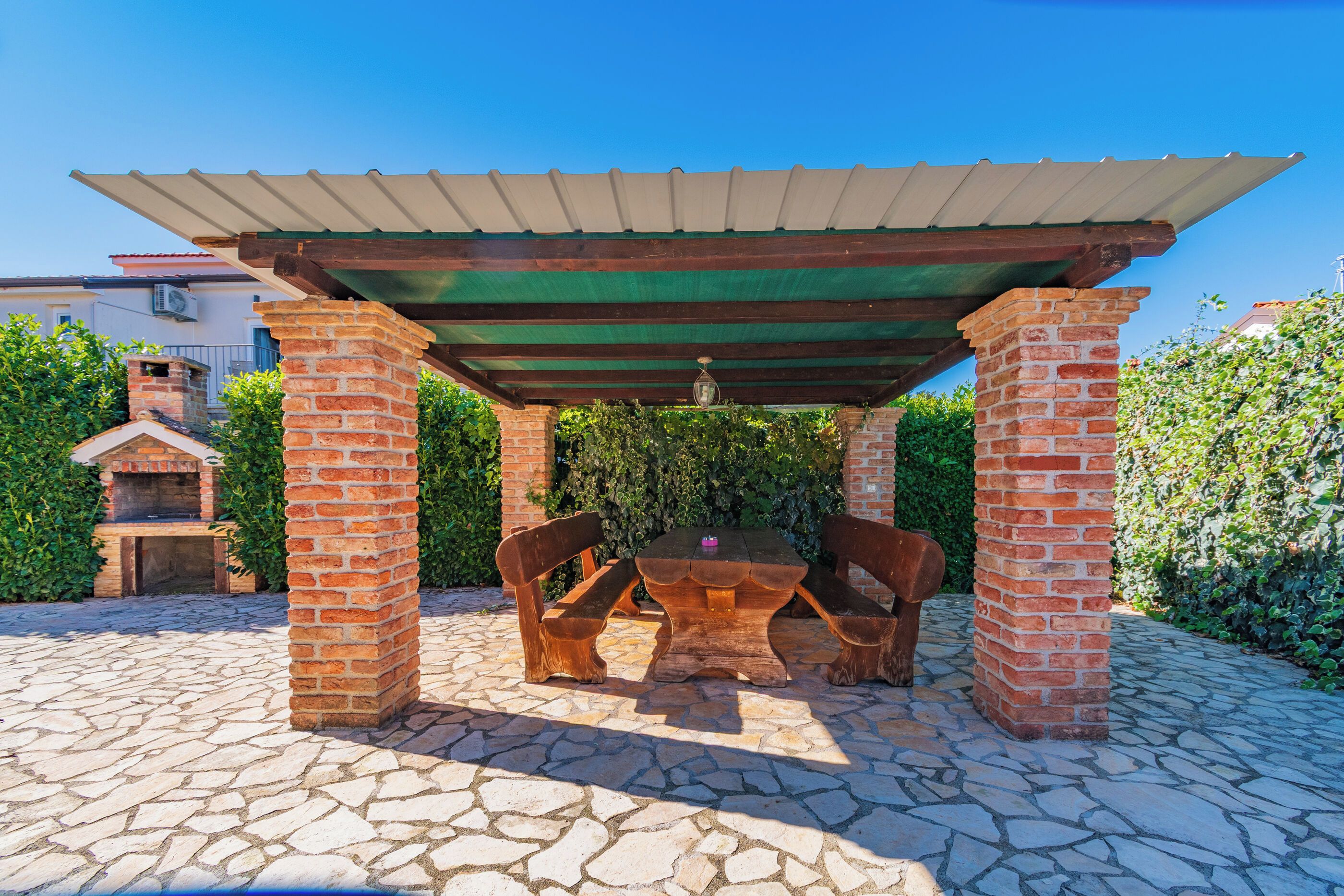 Un patio esterno con tavolo e sedie in mattoni, un pergolato sopra e un camino in laterizio sullo sfondo.