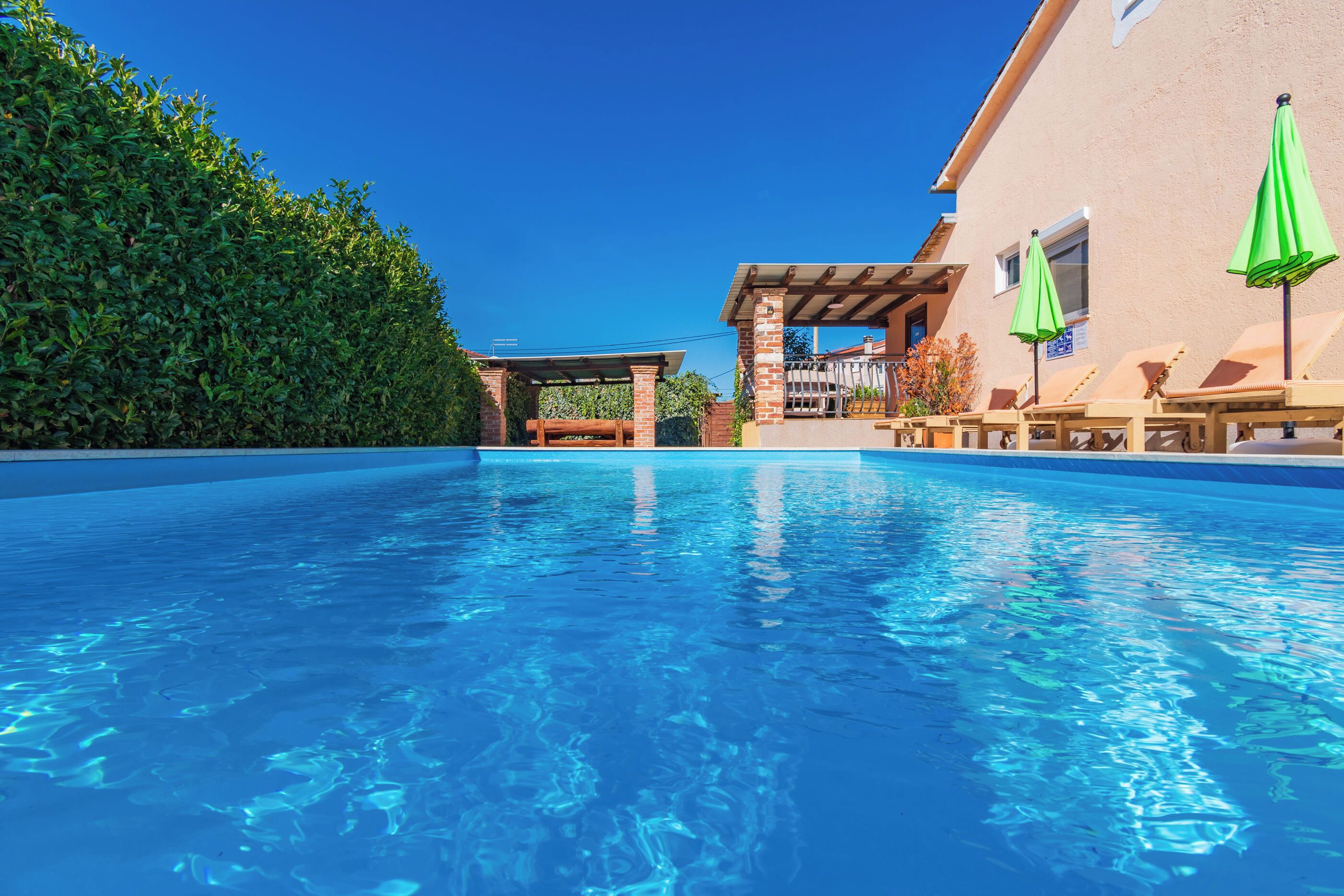 Ein klarer, blauer Pool mit einer Holzterrasse und Liegestühlen, umgeben von einer Hecke und einem Gebäude mit einer Pergola.