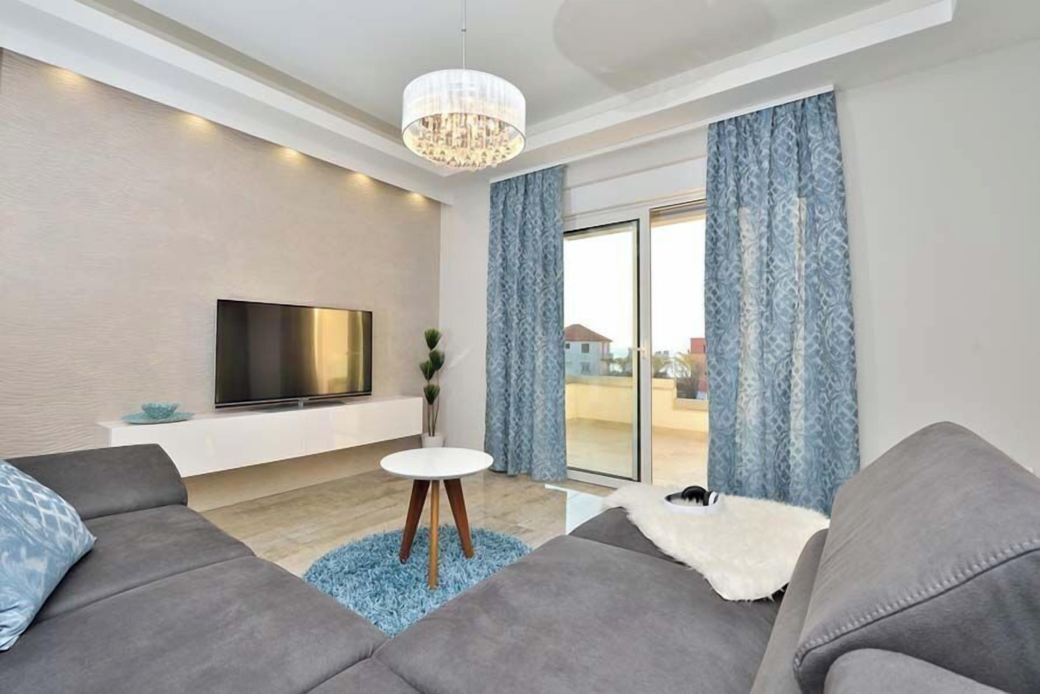 Ein modernes Wohnzimmer mit Flachbildfernseher, grauen Sofas, einem kleinen runden Tisch und einer Kristallleuchtre.