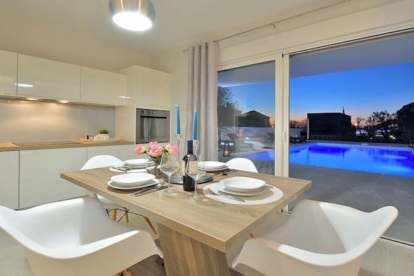 Un comedor moderno con una mesa de madera, sillas blancas y vistas a una piscina al atardecer.