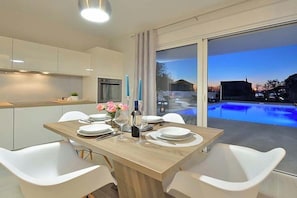 Un comedor moderno con una mesa de madera, sillas blancas y vistas a una piscina al atardecer.