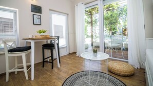 A room with a wooden floor, a white table with a black chair, a round white table, and a wicker ottoman. - Apartments Tellus (Biograd na Moru)