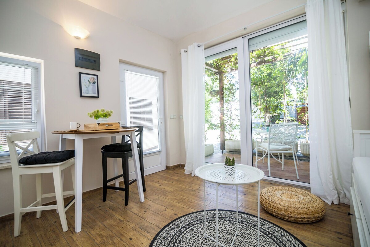 A room with a wooden floor, a white table with a black chair, a round white table, and a wicker ottoman.