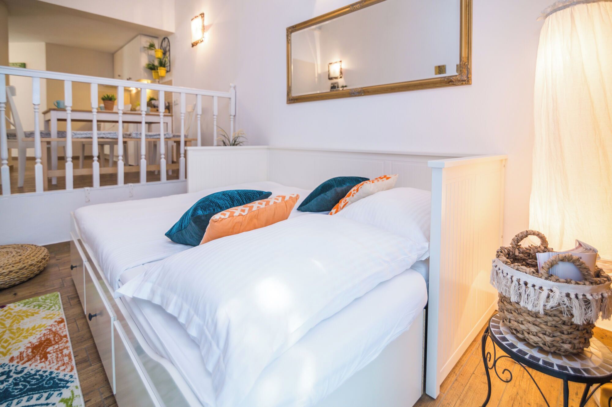 A bedroom with a bed, pillows, a bedside table with a basket, a mirror, and a view of a balcony with a railing.
