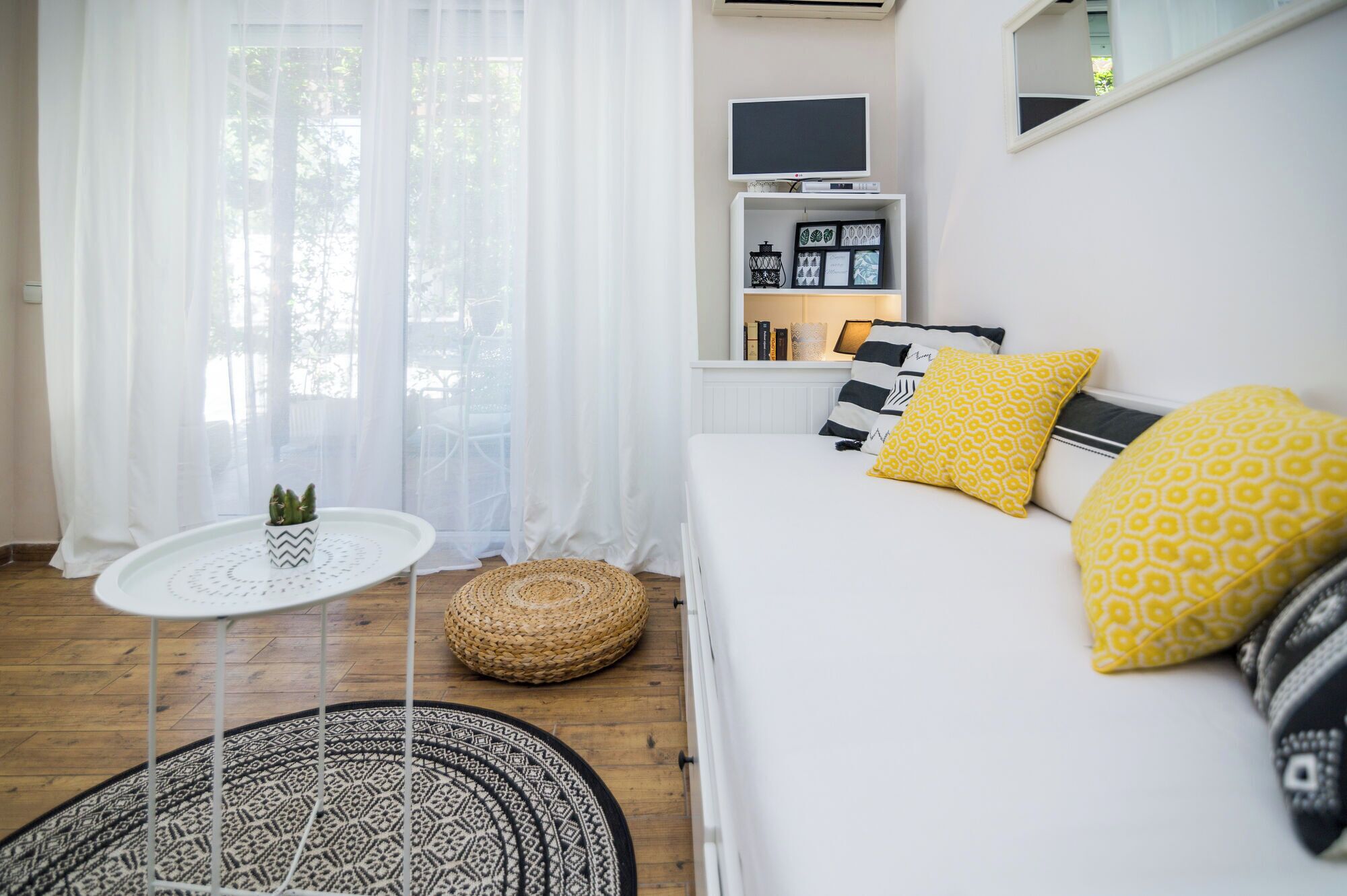 Een slaapkamer met een bed, een klein tafeltje met een plant, een geweven kruk, een televisie op een plank en een raam met dunne gordijnen.