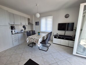 A modern kitchen with white cabinets, a dining table with chairs, a TV, and a clock on the wall. - Apartment Roko (Crikvenica)
