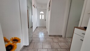 Un couloir avec un sol carrelé, des murs blancs, une porte avec une fenêtre et un petit meuble.