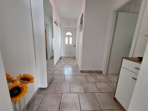 A hallway with a tiled floor, white walls, a door with a window, and a small cabinet. - Apartment Roko (Crikvenica)