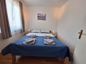 A bedroom with a bed, two bedside tables, a nightstand, a painting on the wall, and a door with a handle. - Apartment Roko (Crikvenica)