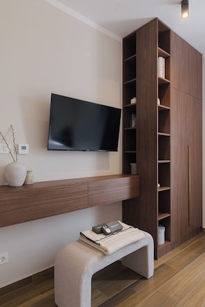 A modern living room with a flat-screen TV mounted on the wall, a wooden bookshelf, a bench with a book, and a small table with a lamp. - Apartments Rooftop Mare Vista (Okrug)