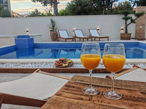 Escena junto a la piscina con dos vasos de zumo de naranja, una mesa de madera y sillones de relax.