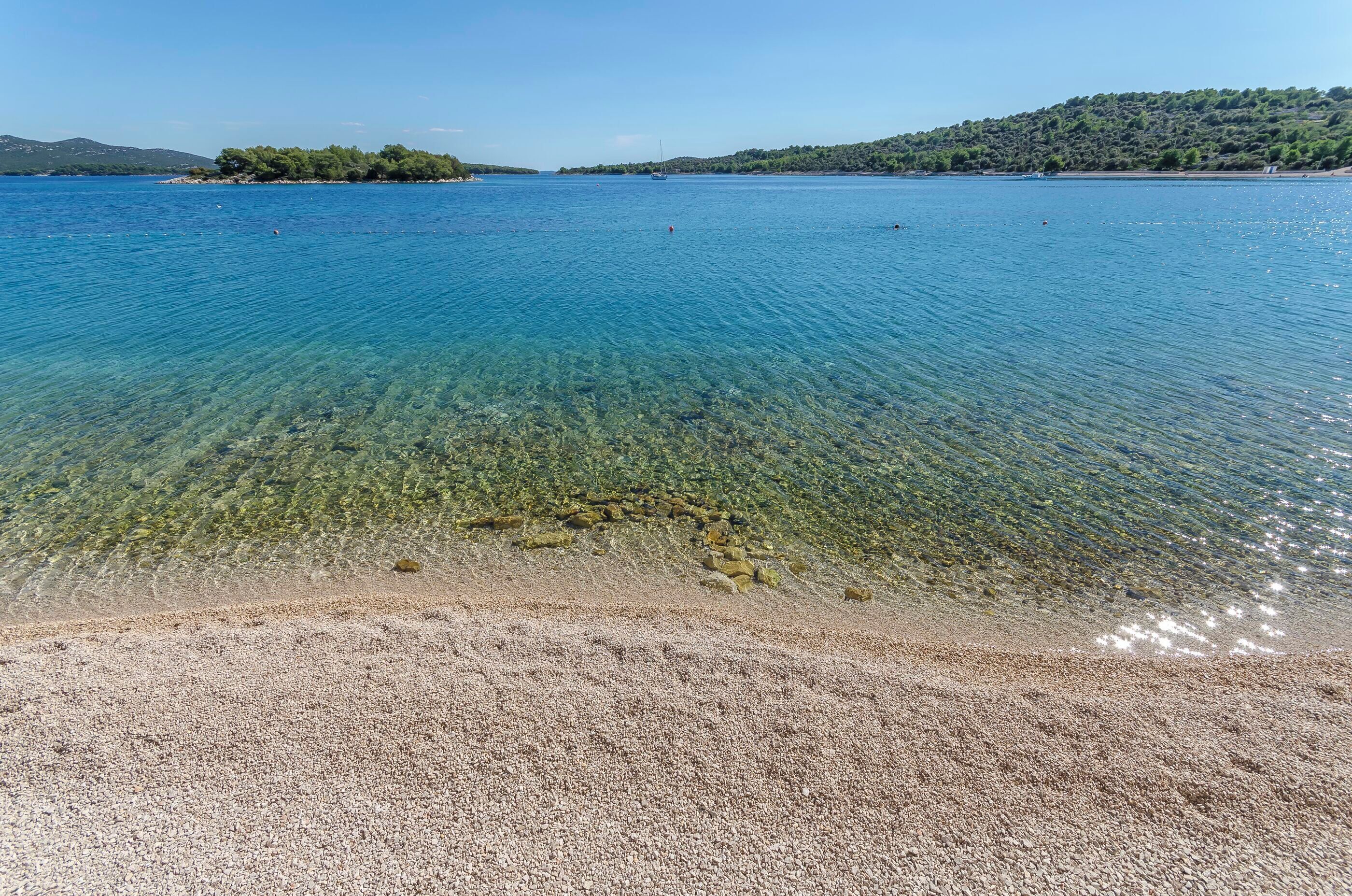 Ein freier Blick auf einen Kiesstrand, der zu einem ruhigen Meer mit einer fernen Insel und Hügeln führt.