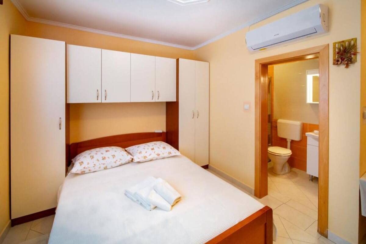A bedroom with a bed, white cabinets, a wooden headboard, an air conditioning unit, and a bathroom with a toilet and sink.