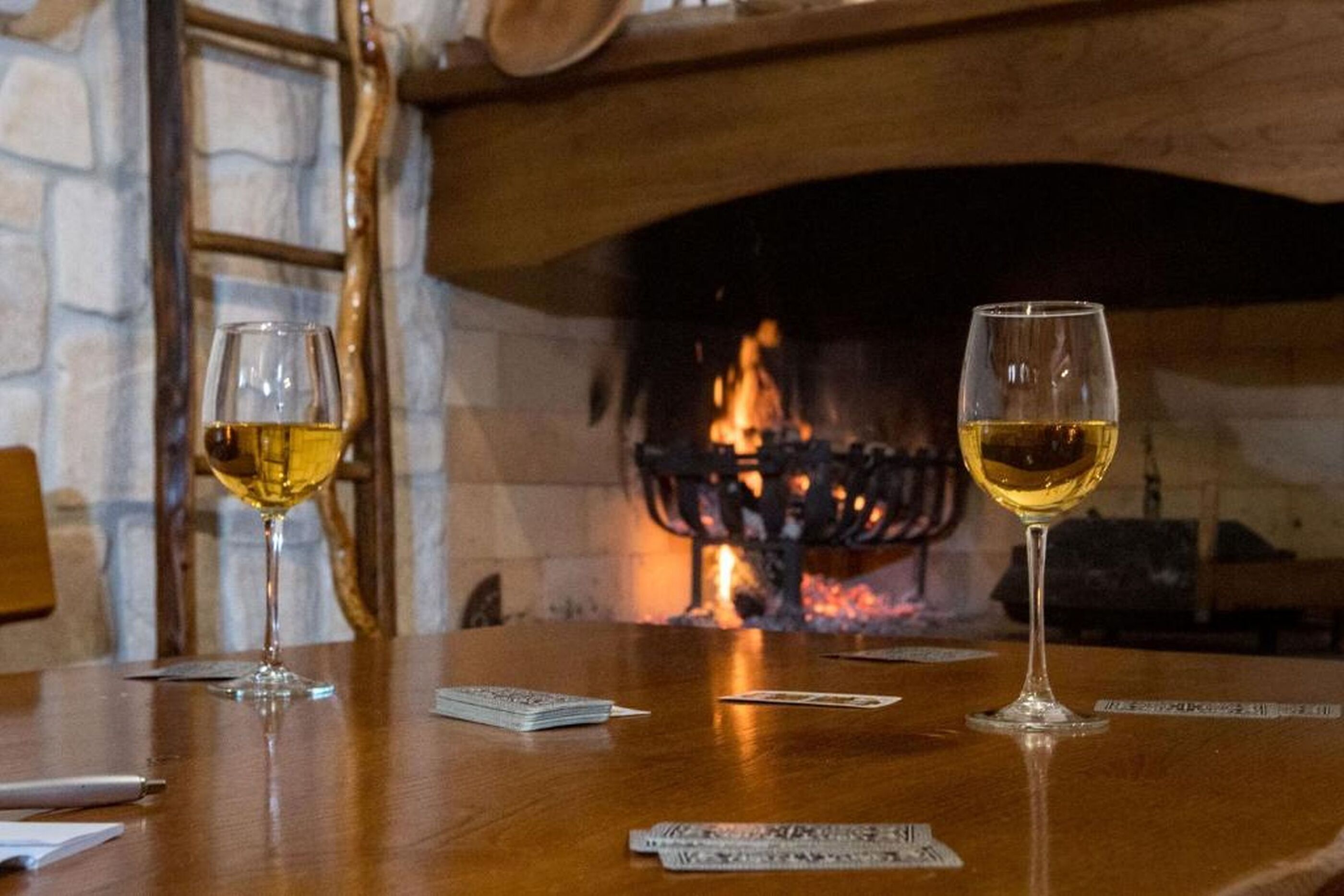 Uma mesa com dois copos de vinho branco e cartas de baralho, com um muro de pedra e lareira ao fundo.