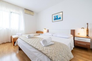 A hotel room with two beds, a large window with curtains, an air conditioning unit, a framed picture on the wall, and a nightstand with a lamp and a giraffe statue. - Apartments Azure View (Makarska)