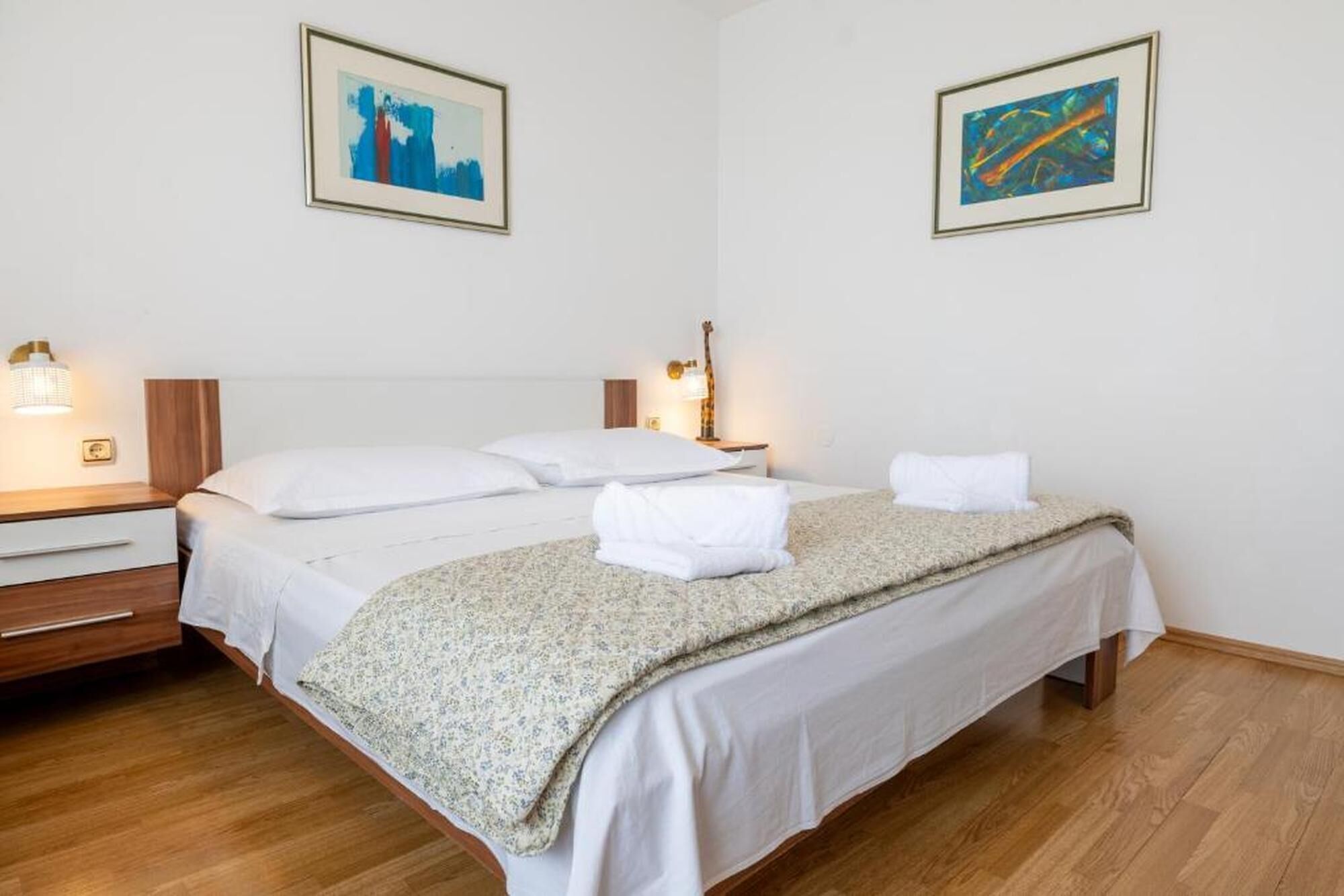 A hotel room with a double bed, two bedside tables, a nightstand, a lamp, and two framed abstract paintings on the wall.