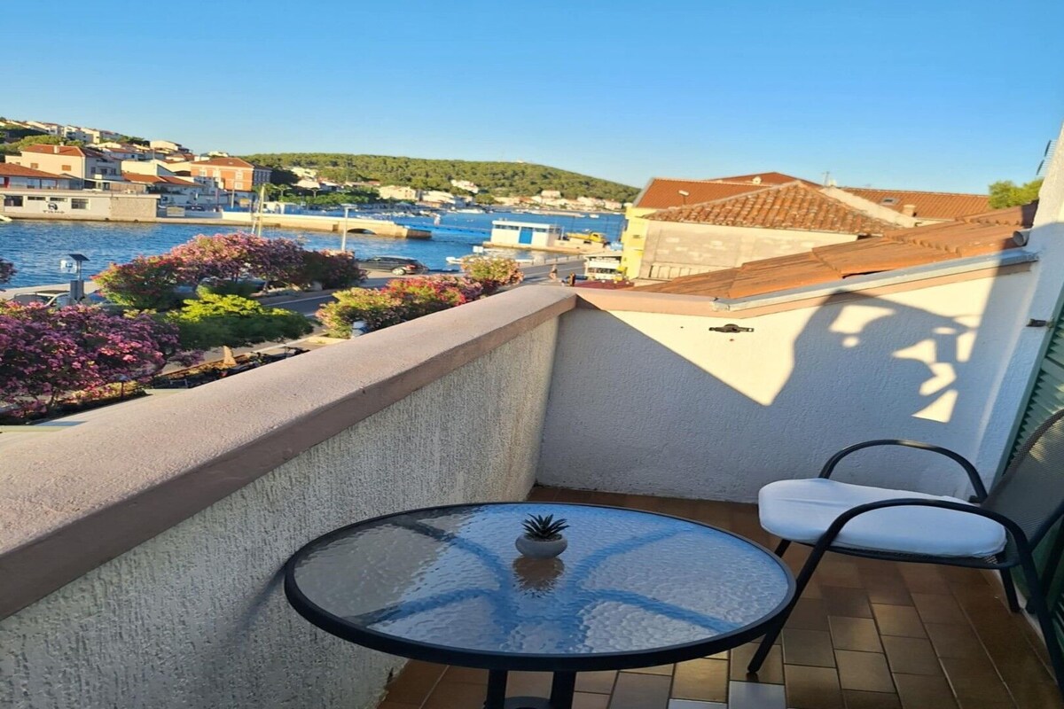 A balcony with a table and chairs, overlooking a coastal town with buildings and a bridge.
