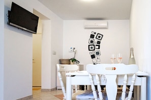 A room with a flat-screen TV mounted on the wall, a white chair with a cushion, a small table with two glasses and a bottle, and an air conditioning unit on the wall. - Apartments Mistral (Lopar)