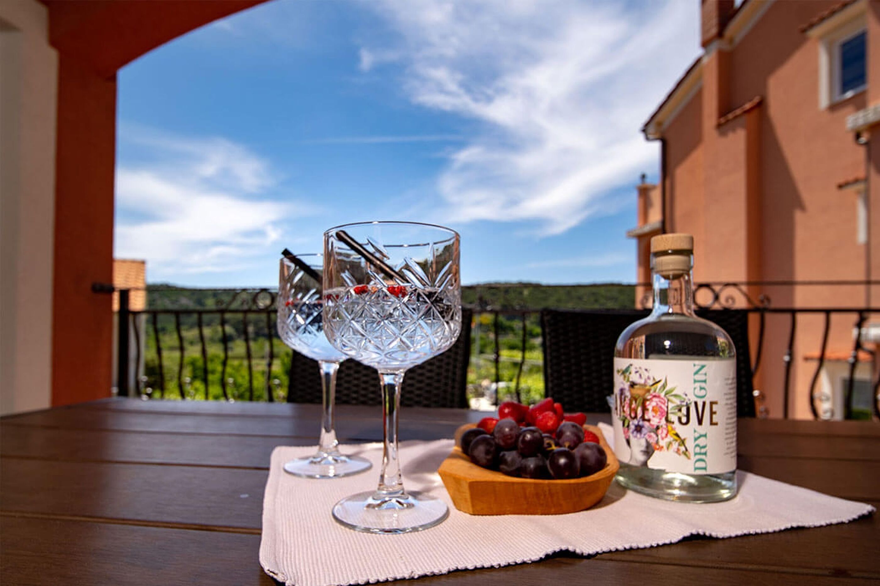 Mesa com dois copos de vidro transparente, uma garrafa de gin e uma tigela de fruta numa varanda com vista para vegetação e um edifício.