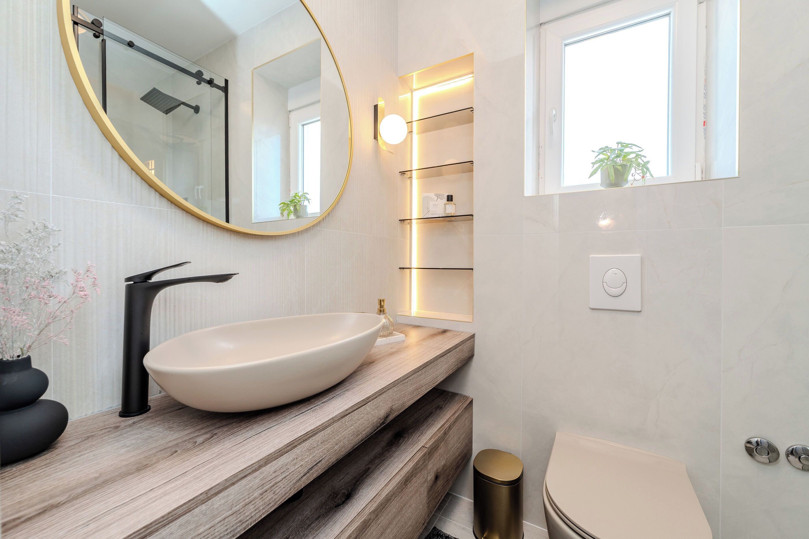 Une salle de bain moderne avec un grand lavabo sur pied, un miroir rond, une cabine de douche en verre et une plante en pot près de la fenêtre.