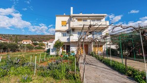 A two-story residential building with a balcony, a garden with various plants, and a clear blue sky. - Apartment Mary (Punat)