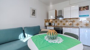 A modern kitchen with white cabinets, a microwave, and a sink. There is a green table with a decorative bowl and a doily. A teal sofa is against the wall. - Apartment Mary (Punat)