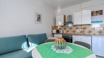 Eine moderne Küche mit weißen Schränken, einer Mikrowelle und einem Spülbecken. Ein grüner Tisch mit einer dekorativen Schale und einer Spitzeneinlage. Ein türkisnes Sofa steht an der Wand.