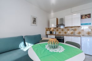 A modern kitchen with white cabinets, a microwave, and a sink. There is a green table with a decorative bowl and a doily. A teal sofa is against the wall. - Apartment Mary (Punat)
