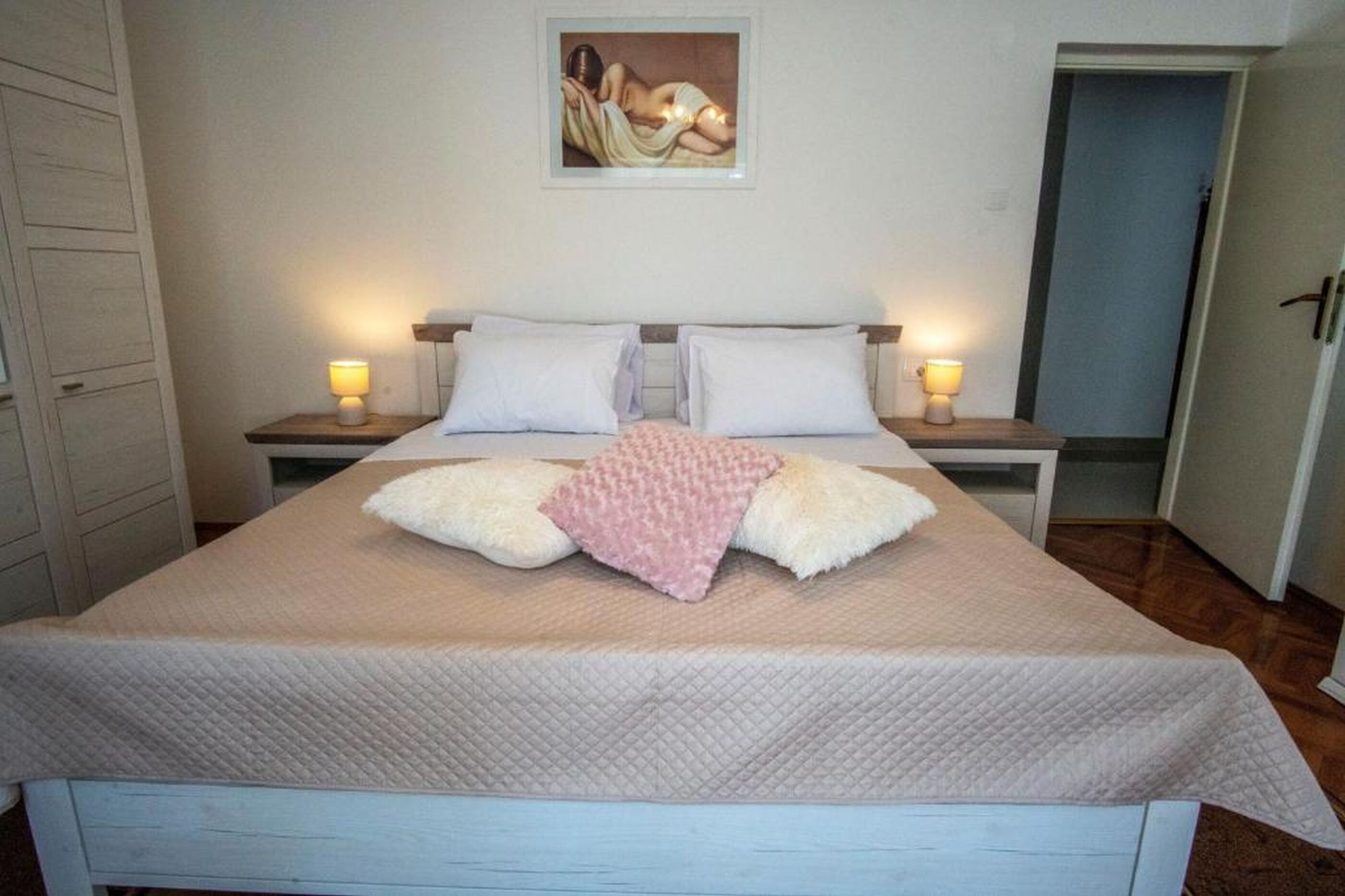 A bed with a quilted cover, two bedside tables with lamps, a painting on the wall, and a door to the right.