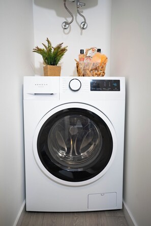 Eine weiße Waschmaschine mit Bedienfeld, ein kleiner Topfpflanze und ein Wäschekorb mit Toilettenartikeln darauf.