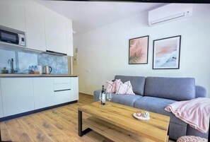 A modern kitchen with white cabinets, a microwave, and a coffee machine. There is a grey sofa with a pink blanket and a coffee table with a bottle and glasses. Two framed pictures are on the wall. - Apartments Sladana (Crikvenica)