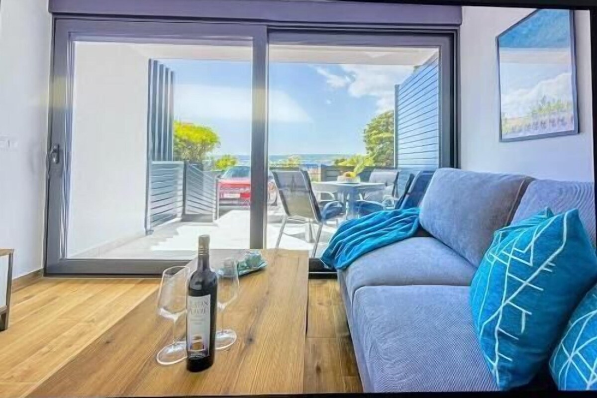 A room with a sofa, a table with a bottle and glasses, and a view through a sliding door to an outdoor area with chairs and a car.