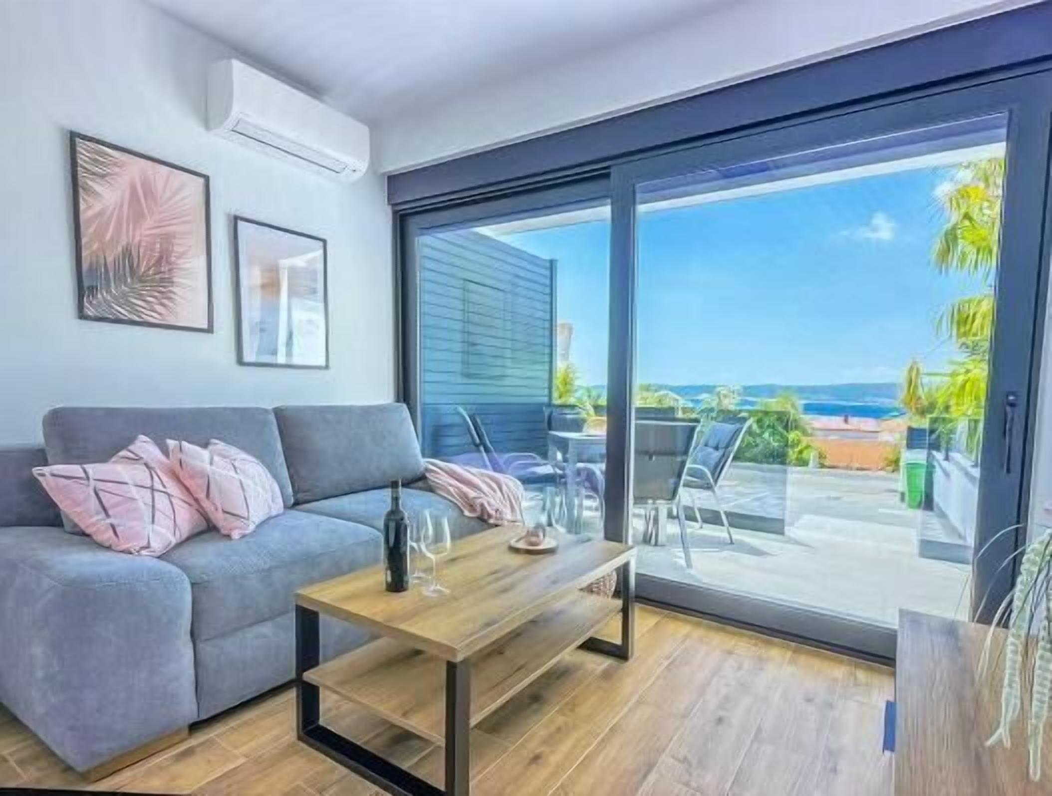 En moderne stue med en grå sofa, et træsofabord og en stor skydedør, der fører ud til en balkon med havudsigt.
