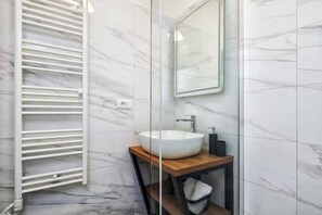 A bathroom with a white marble wall, a wooden vanity, a white sink, a mirror, and a towel rack. - Apartment Tatjana (Split)