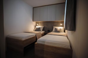 A small, dimly lit room with two wooden beds, a headboard with storage, and a single wall-mounted light. - Mobile Home Aria With Jacuzzi (Vodice)