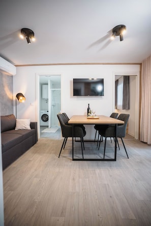 A modern living room with a sofa, a dining table with chairs, a TV mounted on the wall, and a washing machine in the background. - Mobile Home Aria With Jacuzzi (Vodice)
