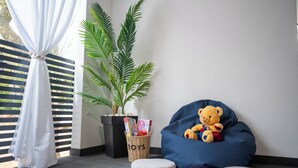Ein Zimmer mit blauem Sitzsack, Teddybär, Topfpflanze, einem Korbwagen mit der Aufschrift 'SPIELZEUG' und weißem Vorhang.
