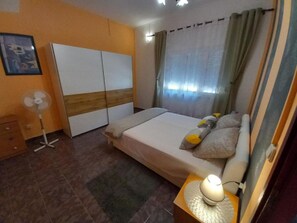 A bedroom with a bed, a bedside table with a lamp, a wardrobe, a standing fan, and a window with curtains. - Apartment Buska (Vir)