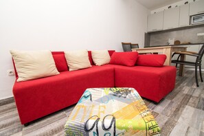 Un sofá rojo con cojines beige y rojos, una mesita con estampado colorido, y una zona de cocina con armarios y un microondas.