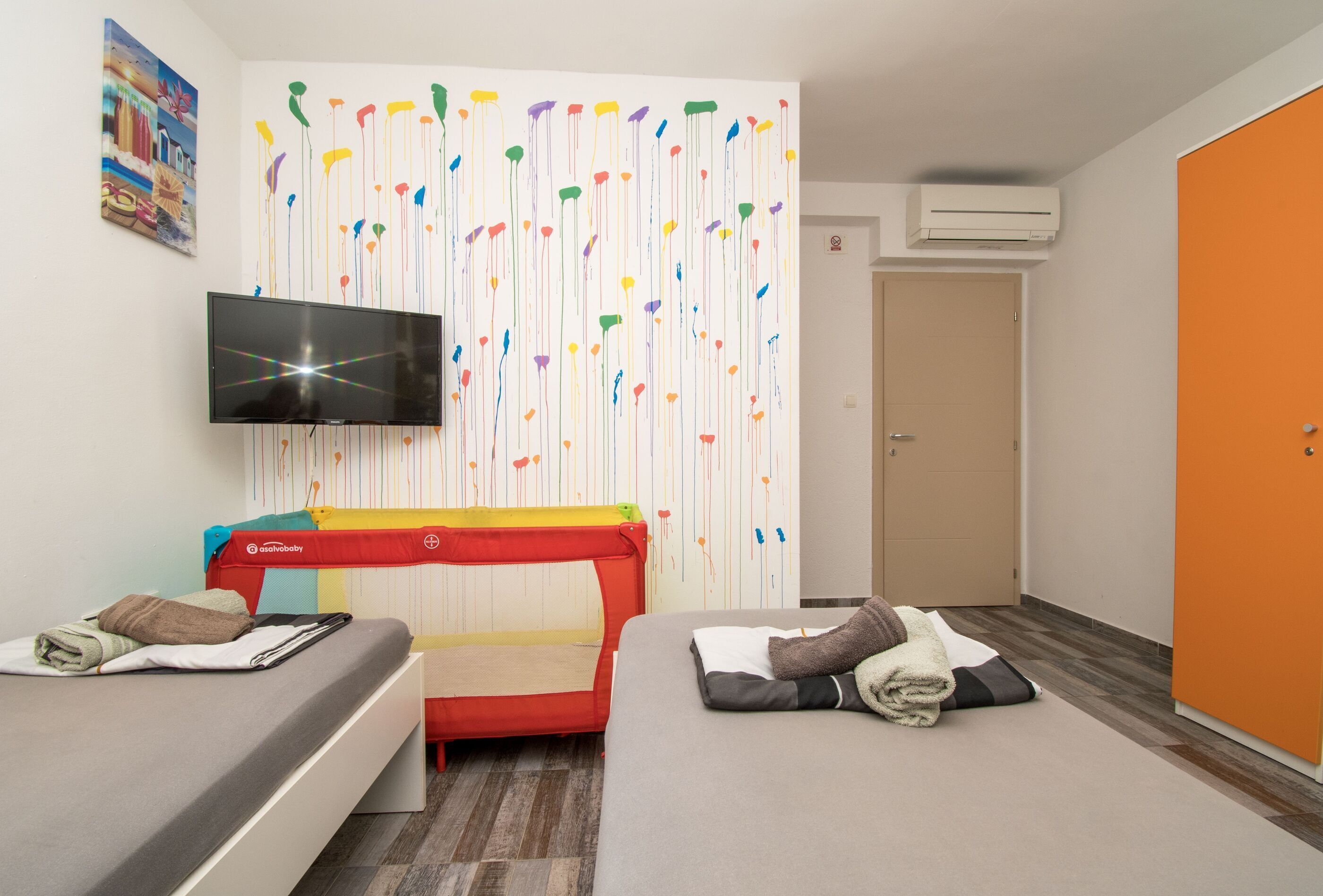 A hotel room with two beds, a television, a colorful wall art, and a door.
