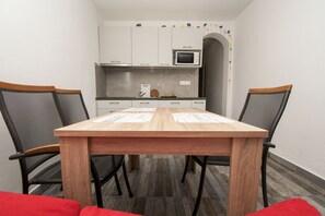 Eine kleine Küche mit weißen Schränken, einer Mikrowelle und einem Spülbecken. Ein Holztisch mit Stühlen und einem roten Kissen ist ebenfalls vorhanden.