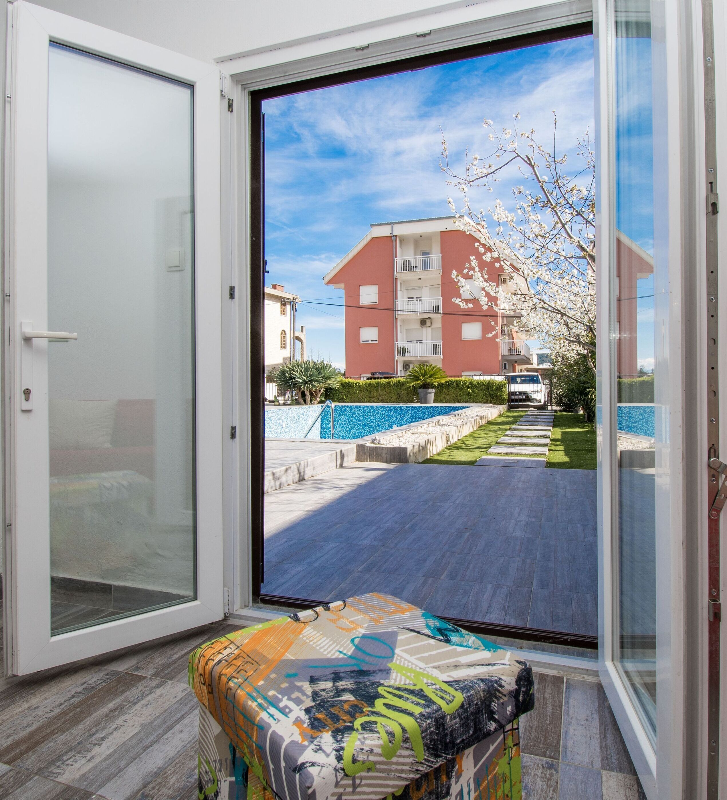 A modern apartment with a sliding glass door leading to a balcony, a patterned ottoman, and a view of a red building and a blue swimming pool.