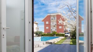 Ein modernes Apartment mit einer Schiebeglastür, die auf einen Balkon führt, einem gemusterten Pouf und Blick auf ein rotes Gebäude sowie einen blauen Swimmingpool.