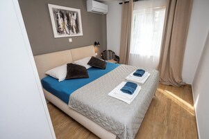 Una cama bien tendida con ropa de cama azul y blanca, un aire acondicionado montado en la pared, un cuadro abstracto enmarcado y una ventana con cortinas.