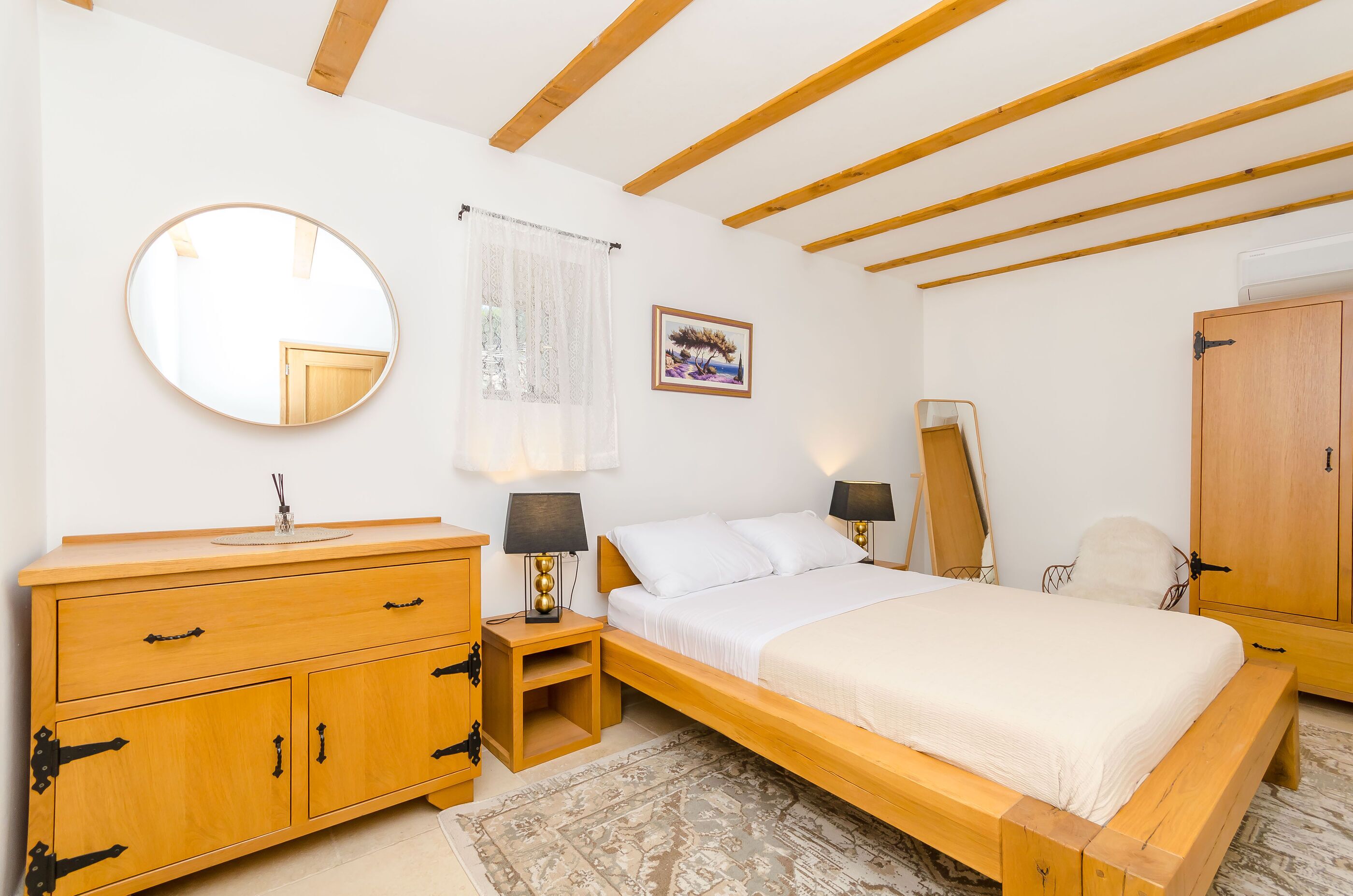 A bedroom with a wooden bed, a wooden dresser, a round mirror, a bedside table with a lamp, and a framed picture on the wall.