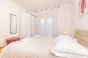 Ein modernes Hotelzimmer mit einem großen Bett, einem Kleiderschrank, einer runden Sitzecke und einem Fenster mit leichtem Vorhang.